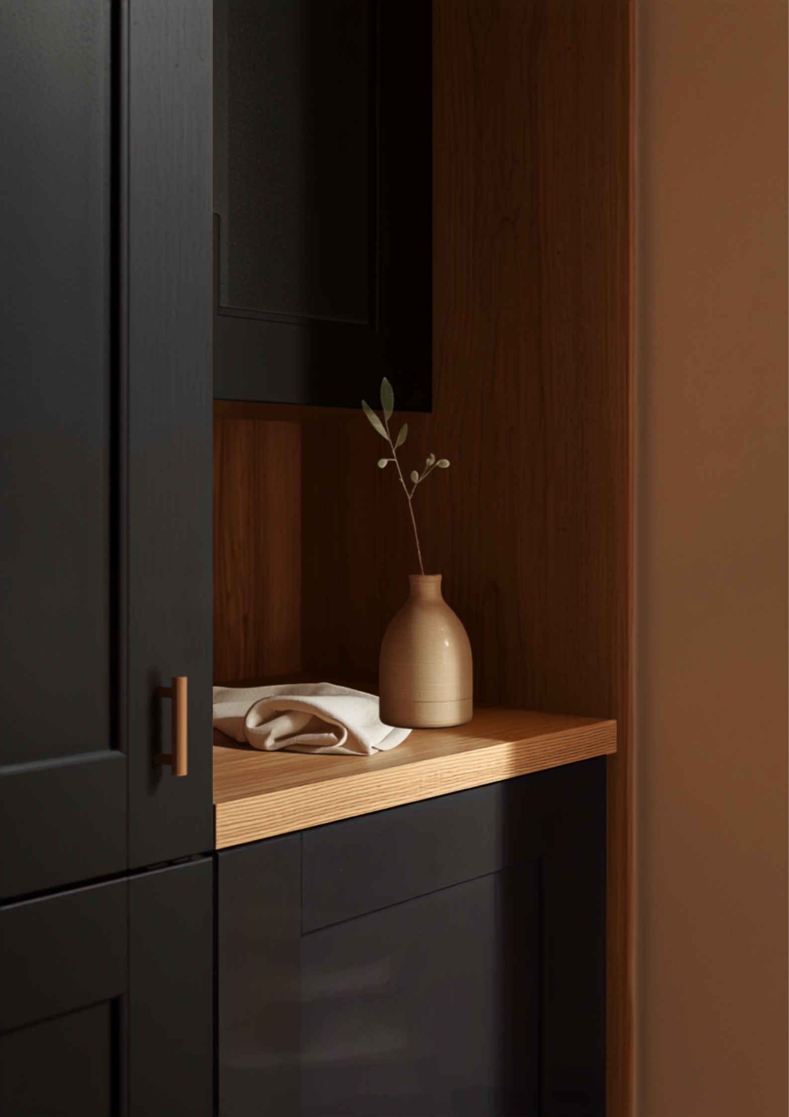 Detail van een matzwarte keukenkast met messing greep naast een warme kleibeige muur en eiken plank met stenen vaasje